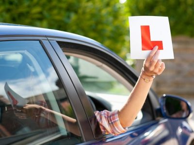 A,Medium,Shot,Of,A,Woman’s,Hand,Holding,A,Learner’s A medium shot of a woman's hand holding a learner's permit sticker out of a car window.